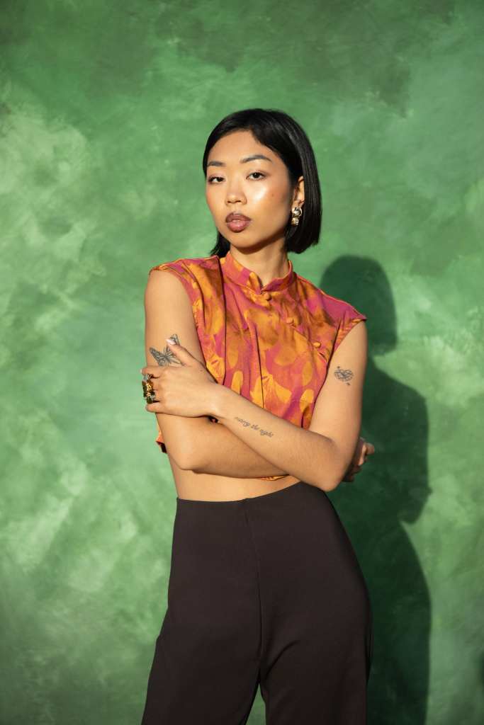 Femme avec un haut coloré et un pantalon noir, posant avec les bras croisés devant un fond vert.